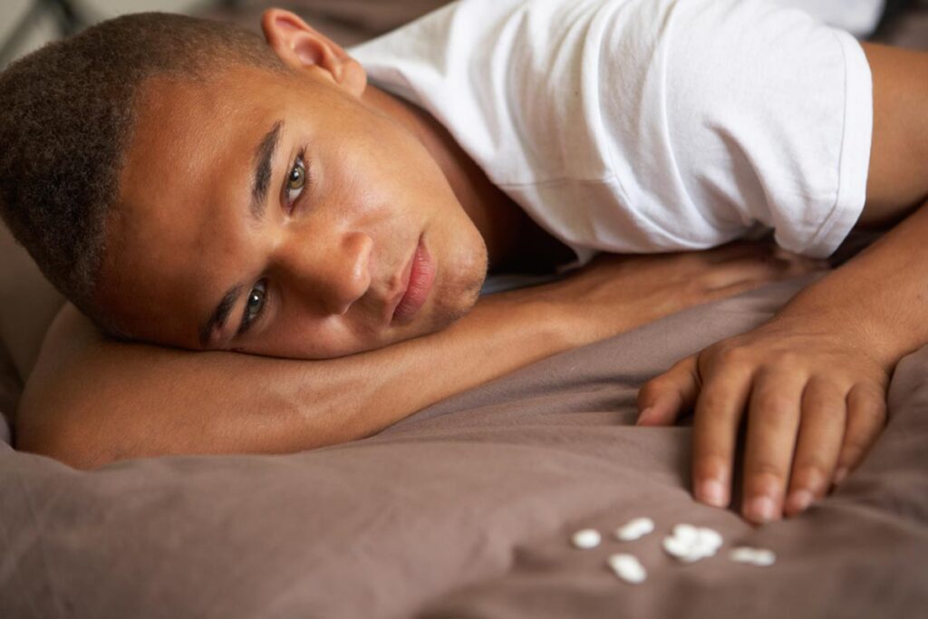lagunashoresrecovery How Normal People End Up Addicted to Opioids photo of Depressed Teenage Boy Lying In Bedroom With Pills